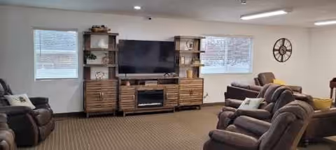 A spacious living room with multiple brown leather recliners and sofas arranged around a large wooden entertainment center holding a flat-screen TV and decorative items. The room has two windows with blinds, a wall clock, and neutral-colored carpet flooring.