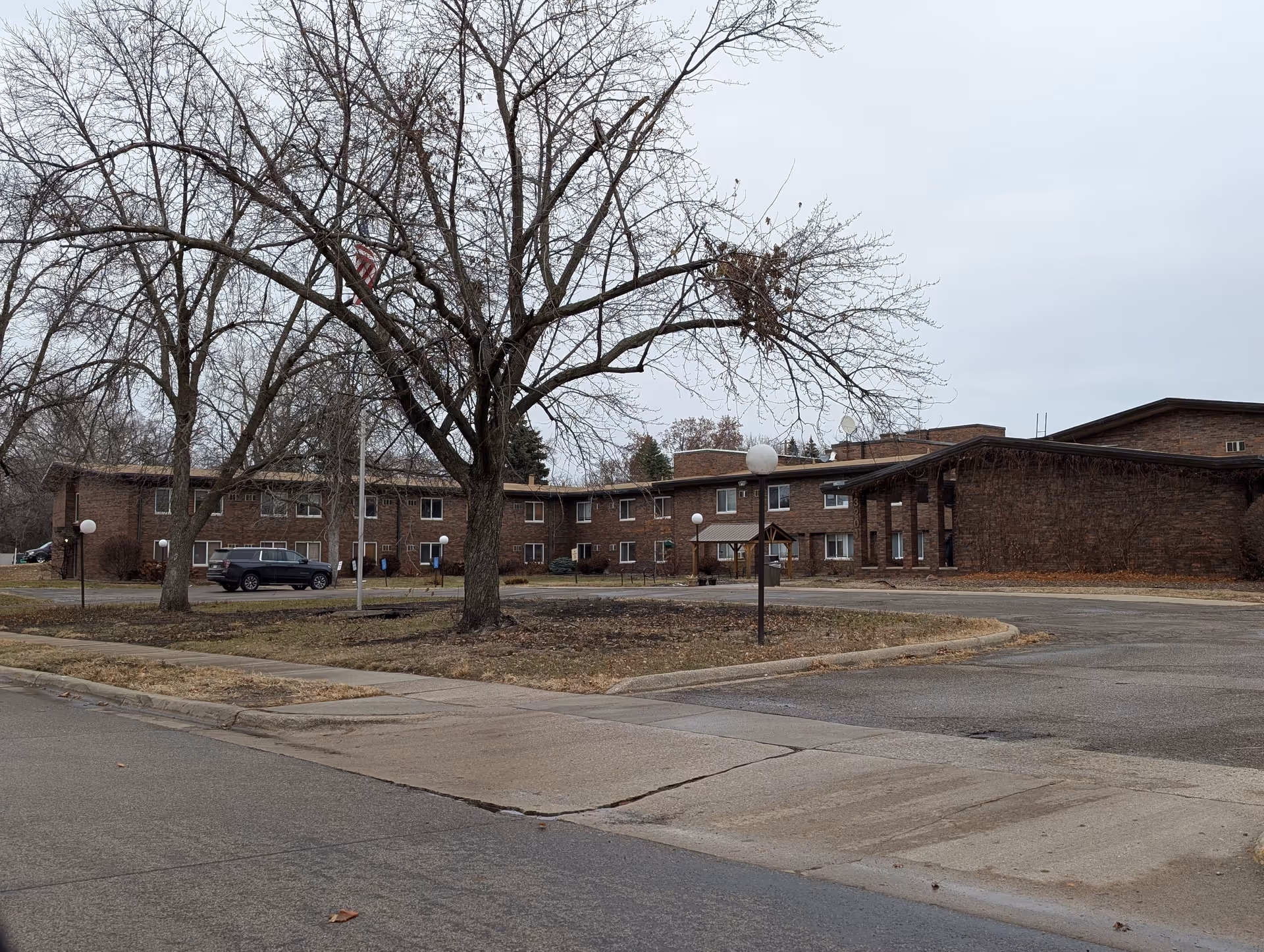 Exterior view of a two-story brick senior living building with leafless trees, a flagpole, and a parking/driveway area.