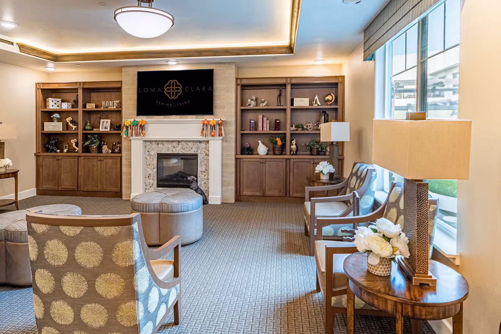 A cozy senior living common area with patterned armchairs, round ottomans, wooden bookshelves filled with decorative items, a fireplace, and a large window letting in natural light. The wall above the fireplace features a screen displaying the Loma Clara Senior Living logo.