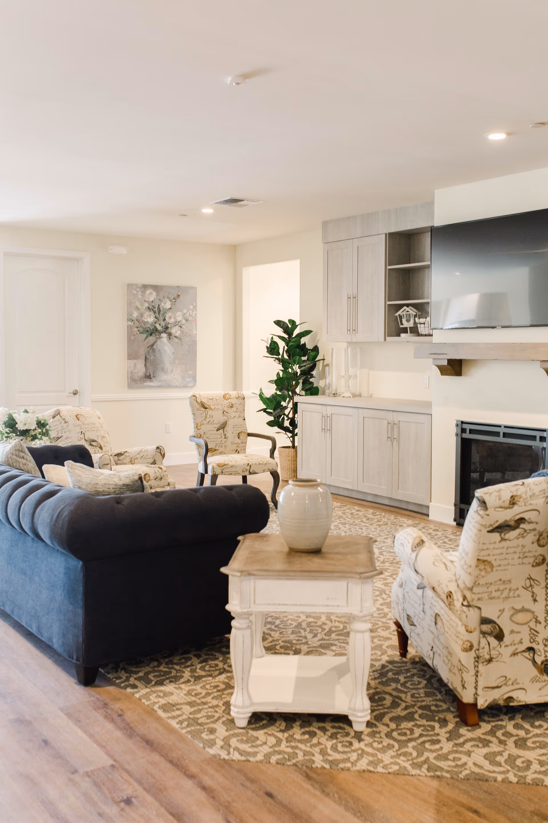 A cozy living room with a dark blue tufted sofa, two patterned armchairs, a white wooden side table with a ceramic vase, a patterned area rug, built-in cabinets, a flat-screen TV mounted above a fireplace, and a potted plant in the corner.