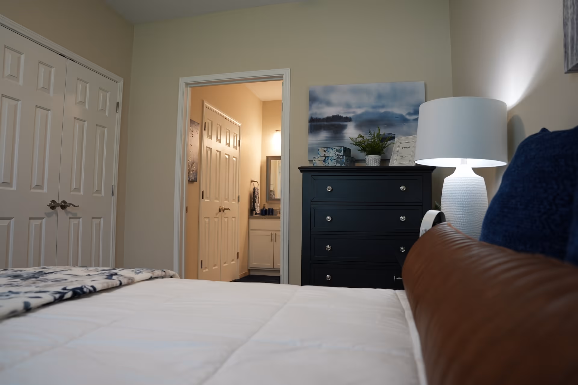 View of a bedroom with a bed in the foreground, a dark dresser with decorative items and a lamp on top, and an open door leading to a bathroom with a sink and mirror visible.