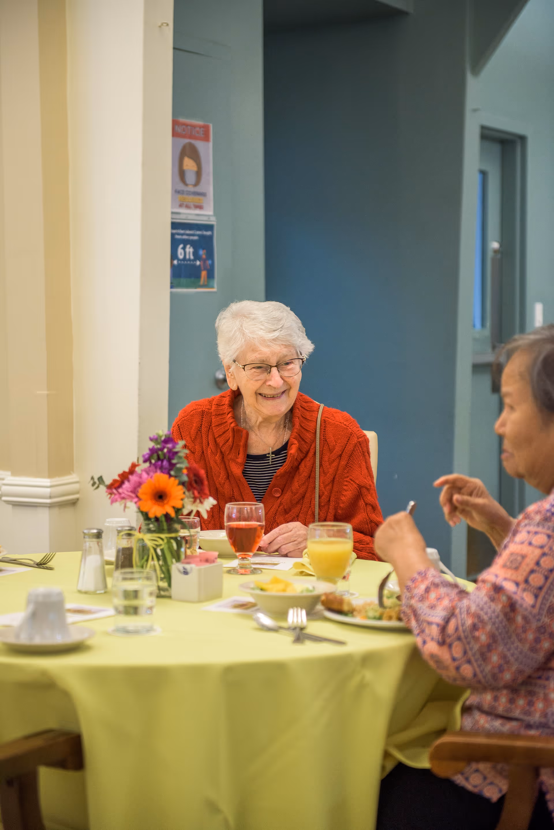Two elderly women seated at a dining table with flowers and drinks, one woman smiling toward the camera.