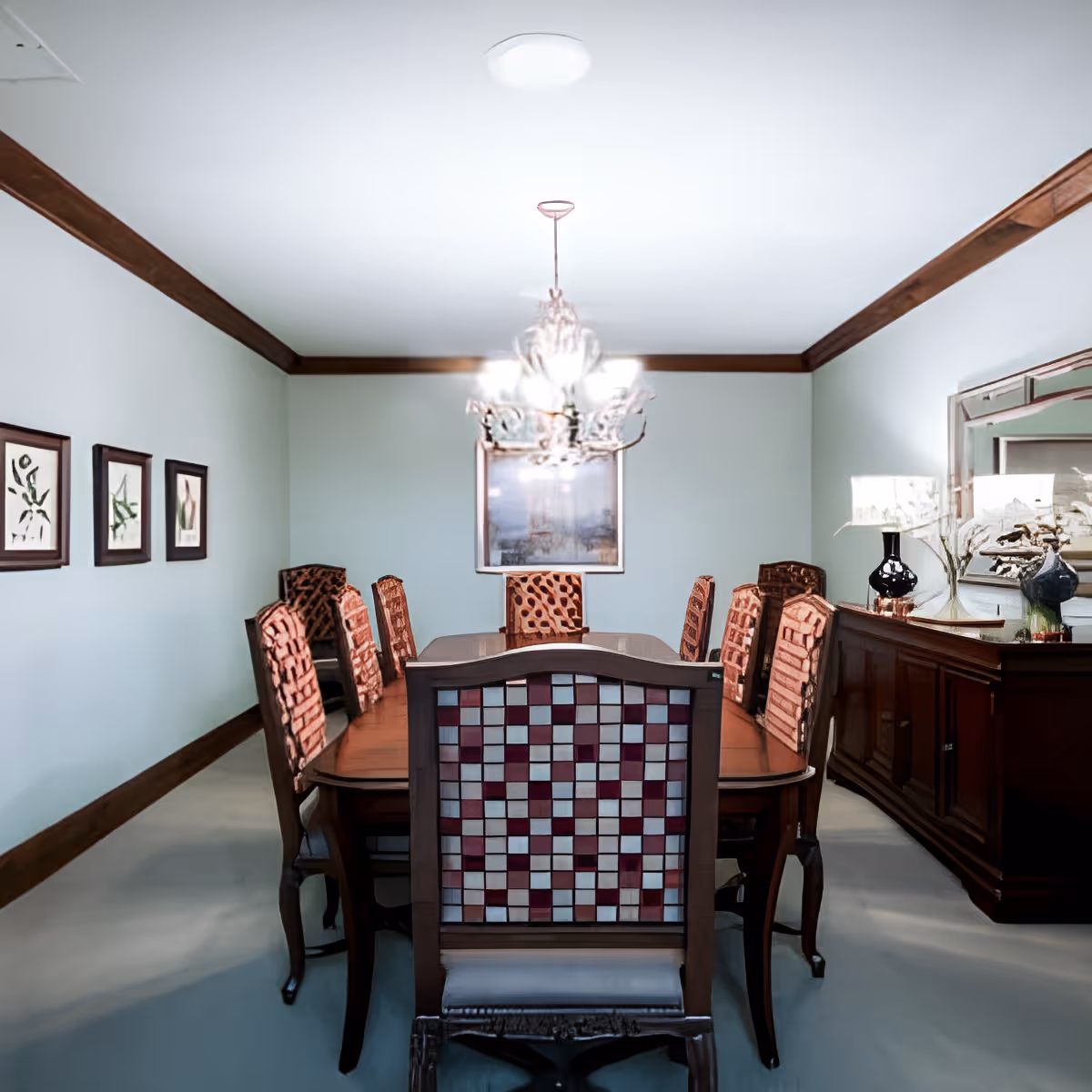 A formal dining room with a long wooden dining table surrounded by eight chairs with patterned upholstery. A chandelier hangs above the table. The room has light-colored walls with wooden trim, three framed botanical prints on one wall, and a wooden sideboard with decorative lamps and a large mirror on the right side.