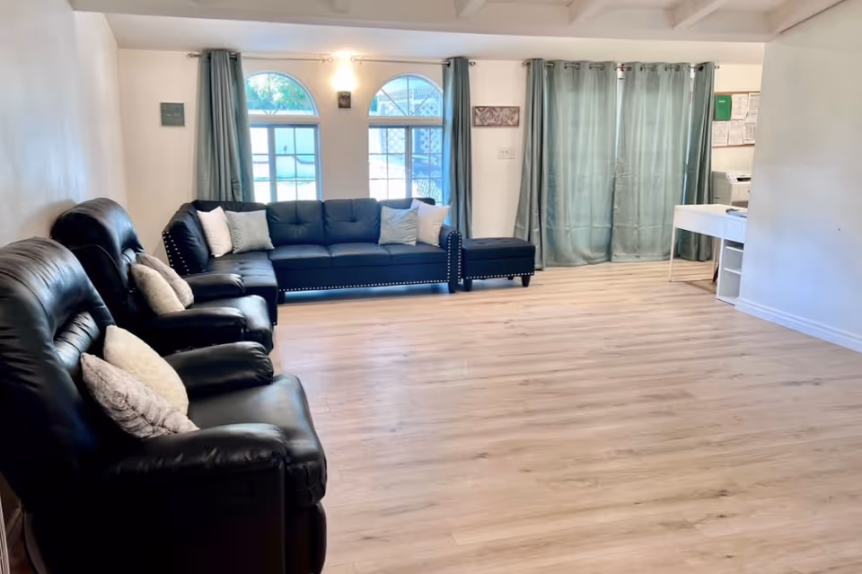 A bright living room with light wood flooring, two black leather recliners with light-colored pillows, a black sectional sofa with white and patterned pillows, two arched windows with teal curtains, and a white desk with office supplies in the corner.