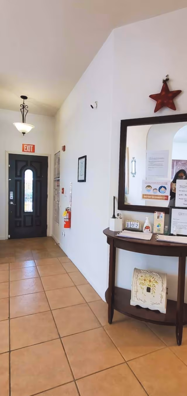 Interior hallway of a facility with a black exit door at the end, a hanging light fixture, and a small wooden table with a mirror above it on the right wall. The table holds various items including hand sanitizer, a tissue box, and informational signs. A red star decoration is mounted above the mirror.