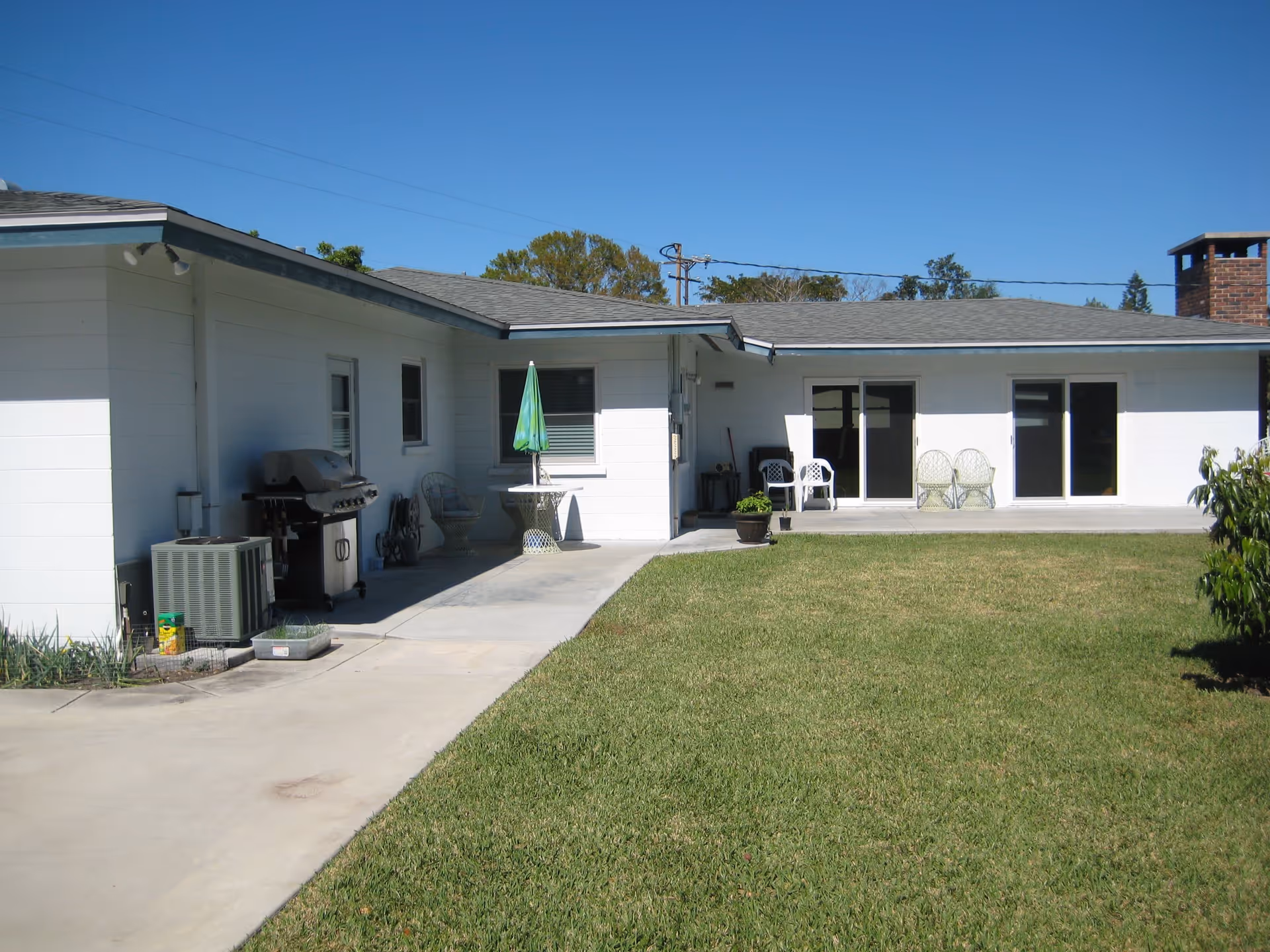 Outdoor view of a single-story white building with a concrete patio and green lawn. There are several chairs, a small table with a green umbrella, a barbecue grill, and some potted plants visible. The sky is clear and blue.