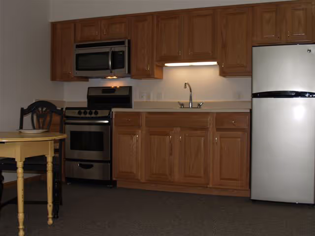 Interior view of a kitchen area with wooden cabinets, a stainless steel microwave above a stove, a sink with a faucet, and a stainless steel refrigerator. A small yellow table with a bowl and two chairs is partially visible on the left side.