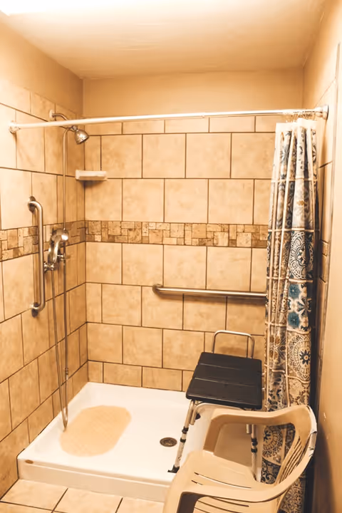 A tiled shower area with beige and brown tiles, equipped with a handheld showerhead, grab bars on the walls, a black shower chair, and a beige plastic chair. A patterned shower curtain is partially visible on the right side.