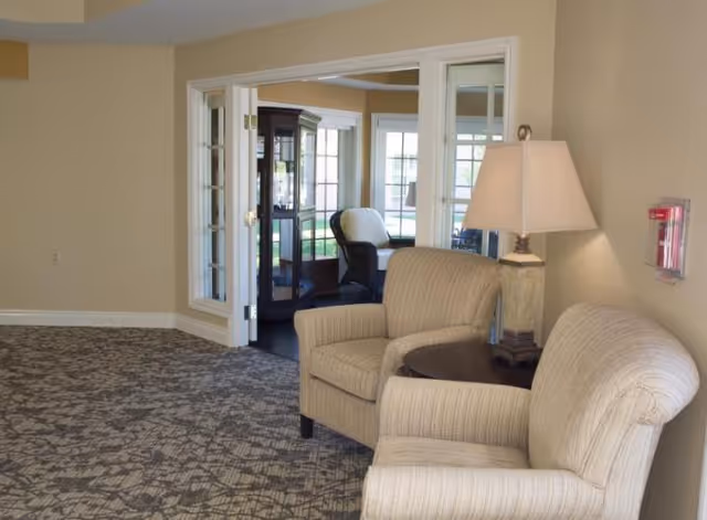 Two upholstered armchairs and a side table with a lamp in a carpeted seating area by glass-paneled double doors leading to a sunroom.