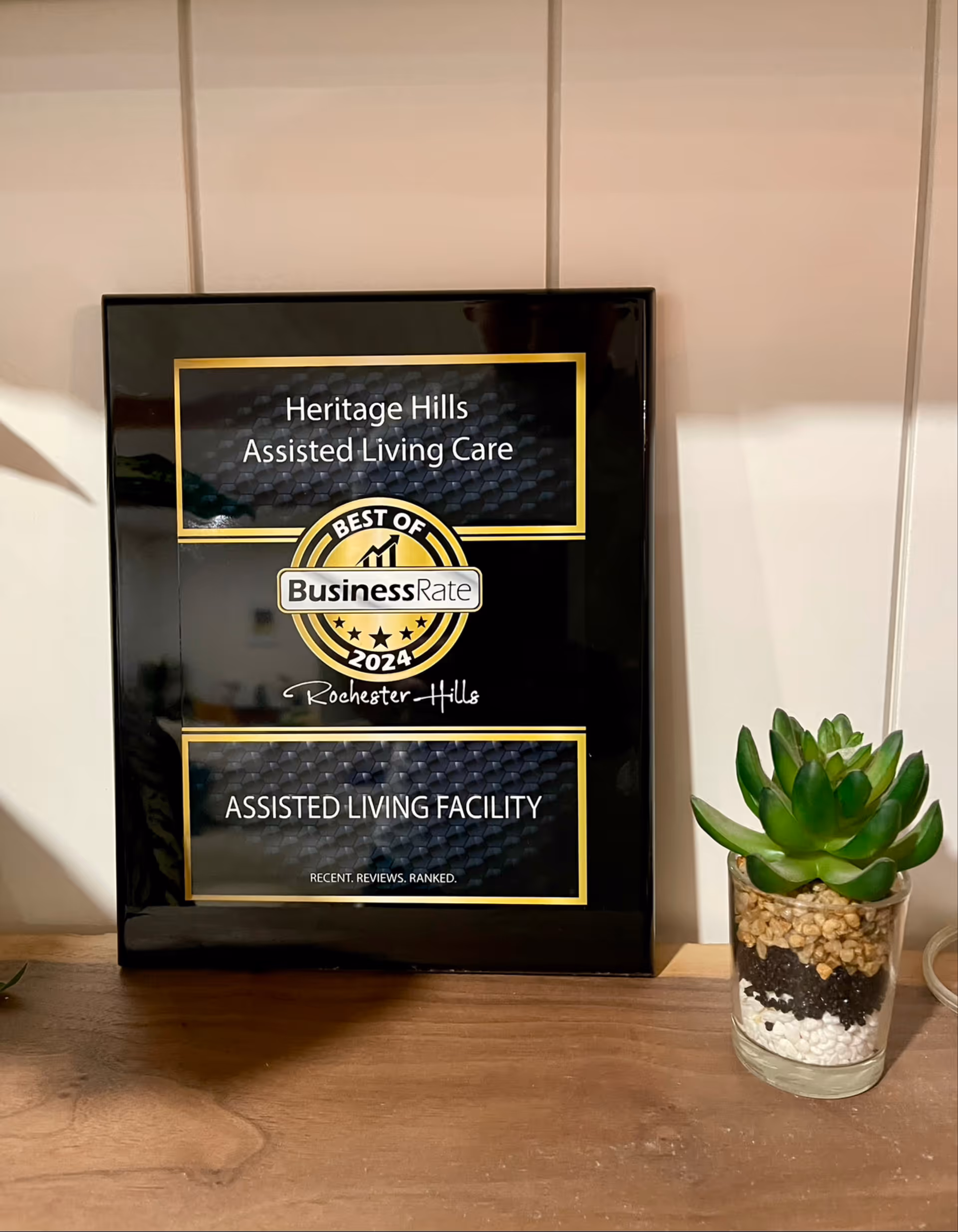 A black and gold framed award plaque for Heritage Hills Assisted Living Care, labeled Best of Business Rate 2024 Rochester Hills, placed on a wooden surface next to a small glass container with a layered succulent plant.