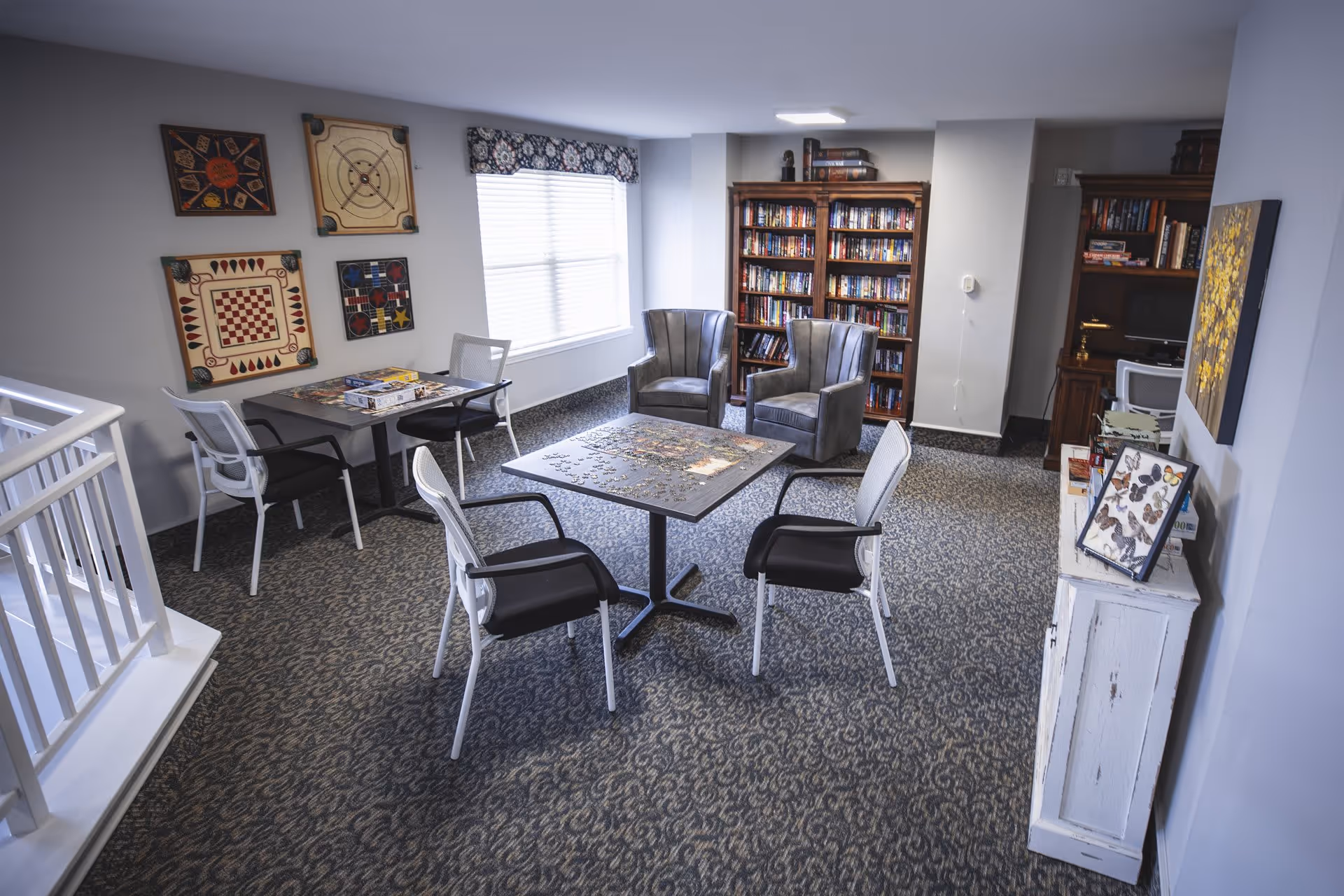 A cozy common area with a carpeted floor featuring a patterned design. There are two tables with chairs around them; one table has a partially completed jigsaw puzzle, and the other has board games. On the walls, there are framed game boards and a window with a decorative valance. In the background, there are two gray armchairs in front of a wooden bookshelf filled with books. A small white cabinet with framed butterfly artwork and other items is visible on the right side of the image.