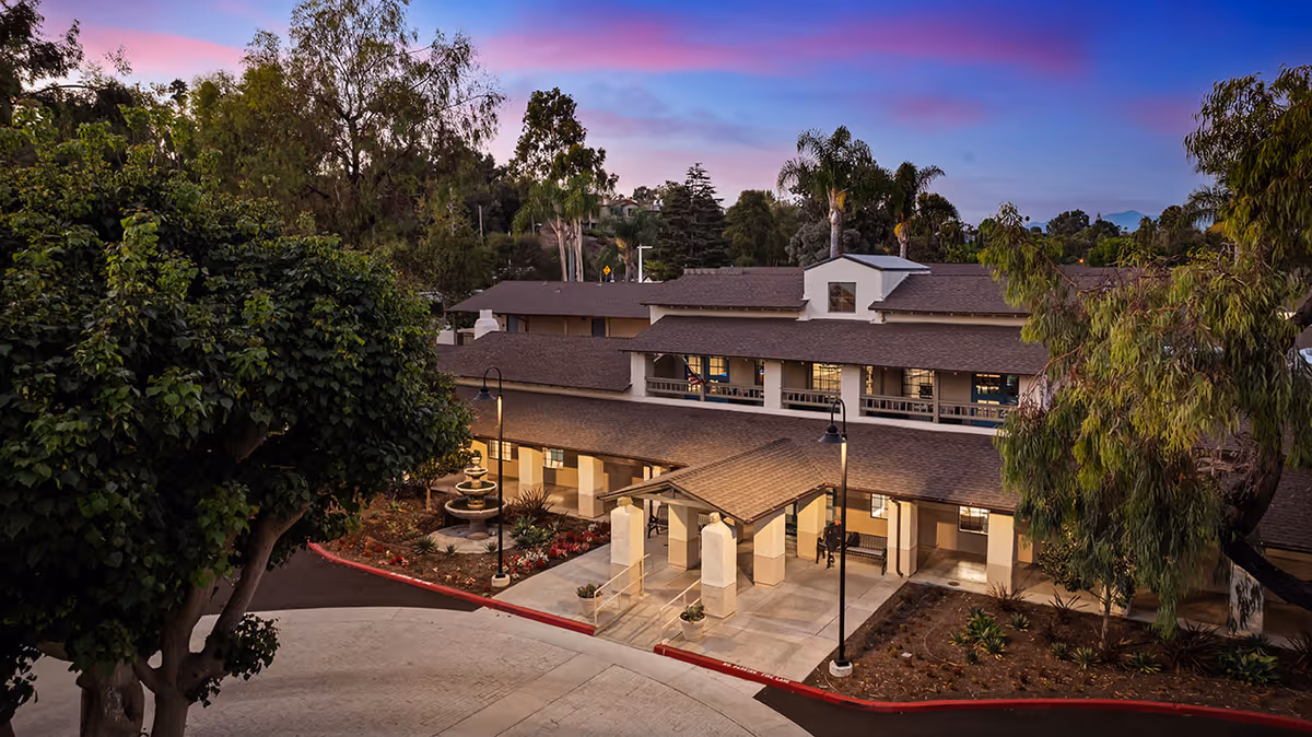 Exterior view of a senior living facility building at dusk with a covered entrance, surrounded by trees and landscaping under a colorful sky.