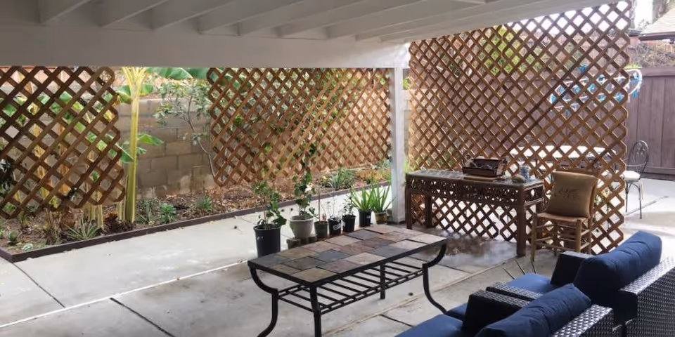 Covered outdoor patio area with lattice wood panels, potted plants, a stone-tiled coffee table, a blue cushioned sofa, a wooden chair with a cushion, and a decorative wooden table. The patio overlooks a small garden bed with plants and a brick wall.