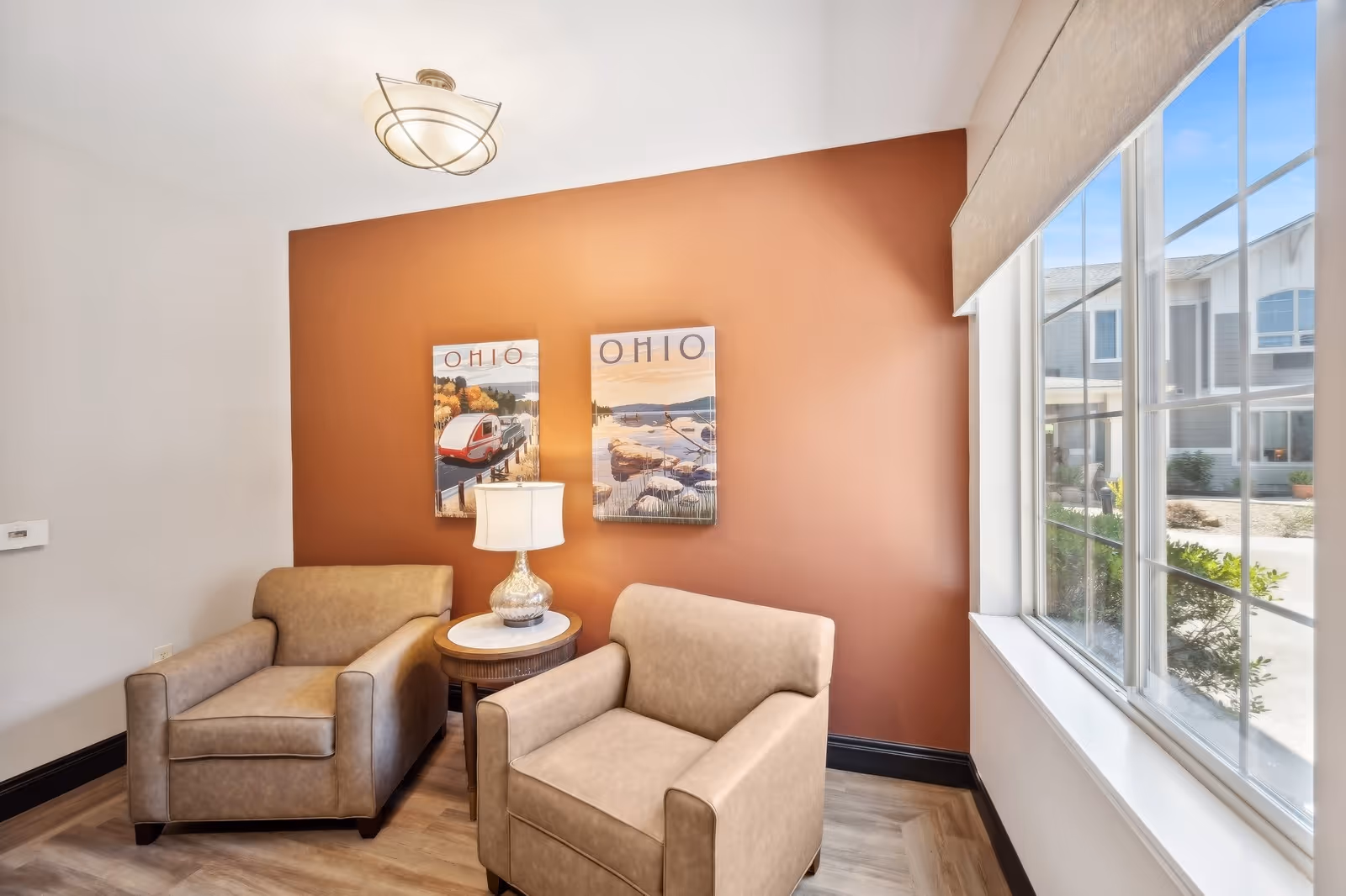 A cozy seating area with two beige armchairs separated by a round wooden side table with a lamp on it. The wall behind the chairs is painted a warm terracotta color and decorated with two framed pictures labeled 'OHIO' depicting scenic landscapes. A large window on the right side lets in natural light and offers a view of the building exterior and courtyard.