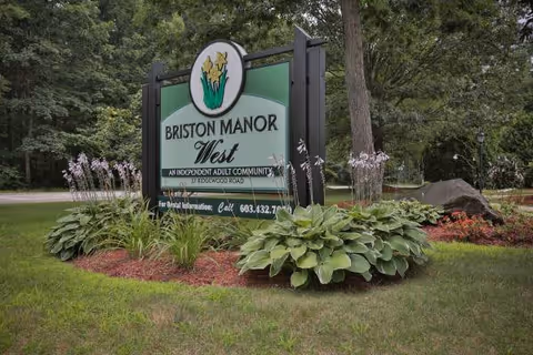 Outdoor sign for Briston Manor West, an independent adult community, surrounded by green plants and flowers with trees in the background.