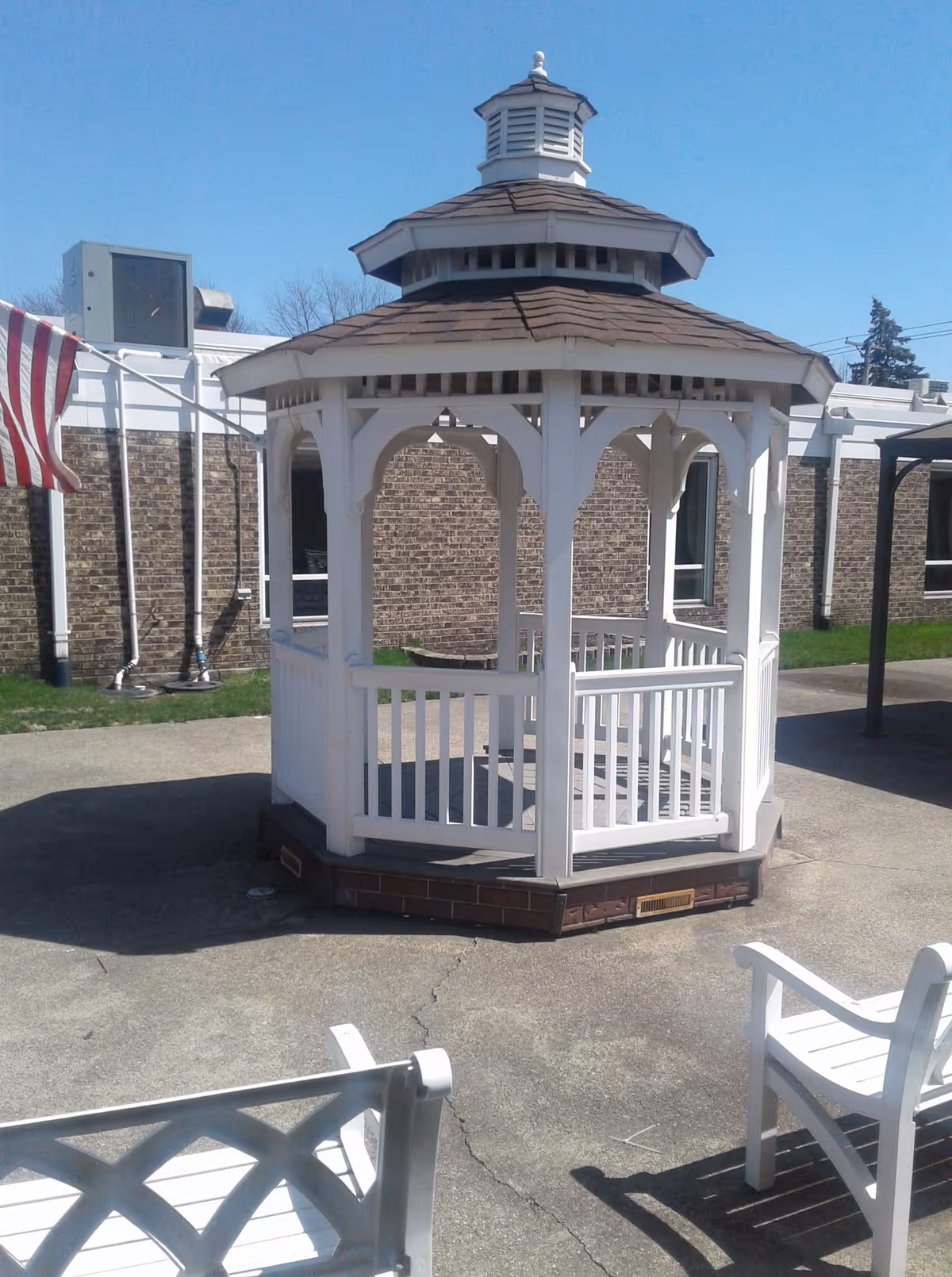 A white wooden gazebo with a brown shingled roof situated on a concrete patio area outside a brick building. There are white benches around the gazebo and an American flag partially visible on the left side. The sky is clear and blue.