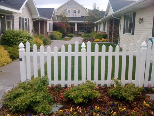 White picket fence in front of a landscaped courtyard with walkways and adjacent assisted living buildings.