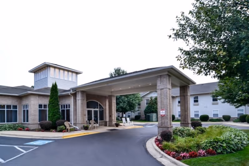 Exterior view of Cedarhurst Senior Living of Edison Lakes showing the main entrance with a covered driveway, surrounded by landscaped greenery and trees, with a clear sky above.