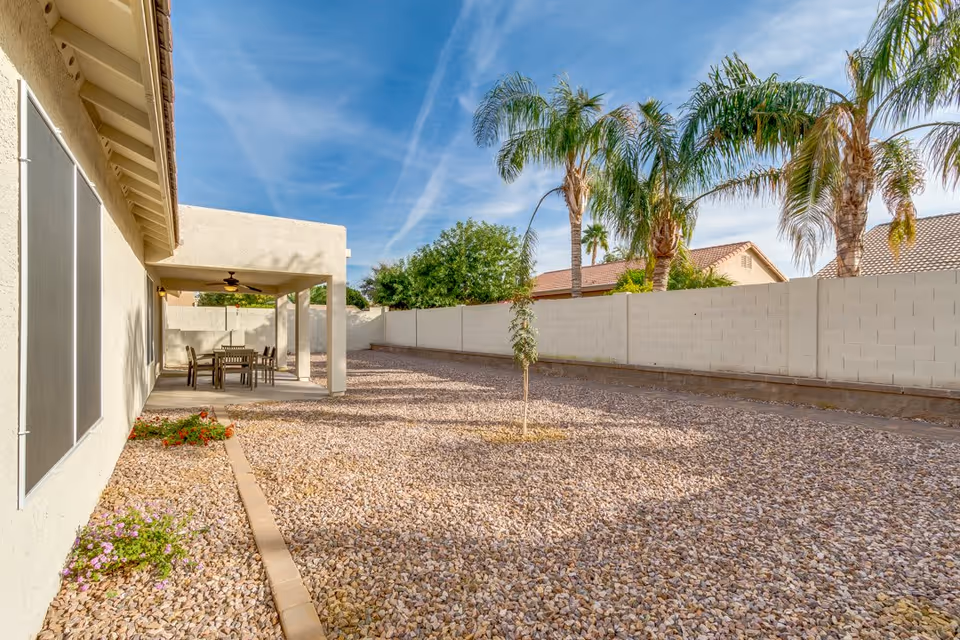 Outdoor area with a covered patio featuring a table and chairs, surrounded by a gravel yard with a small tree and some plants near the building. Palm trees and a white block wall fence are visible under a blue sky with some clouds.