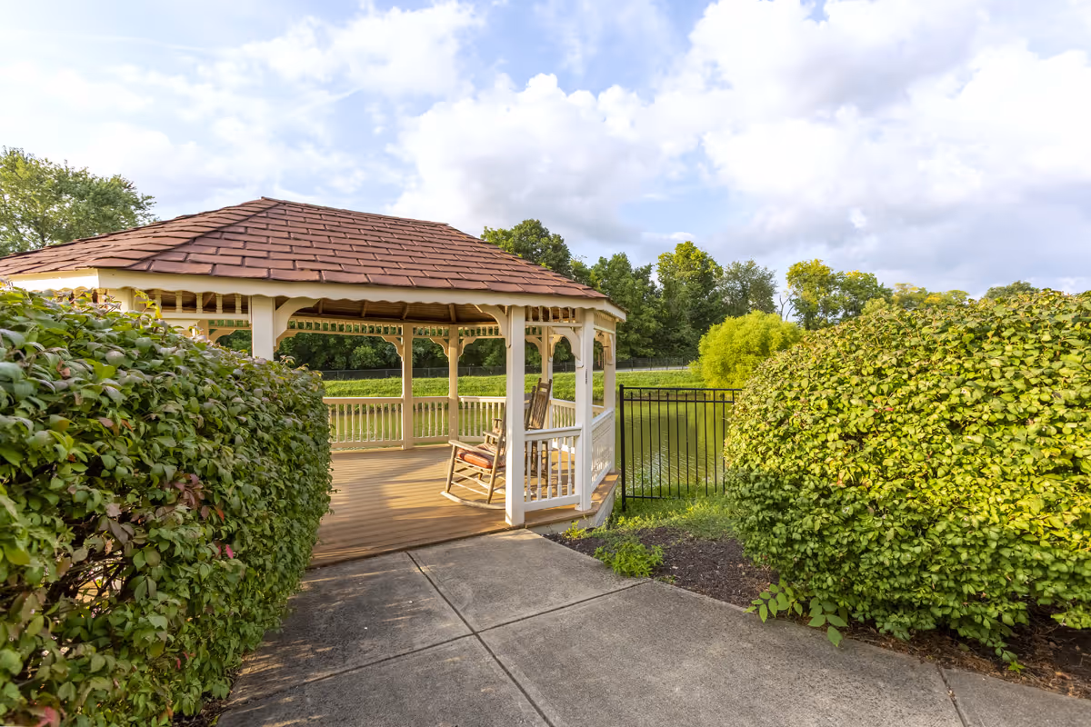 White gazebo with rocking chairs beside a pond surrounded by hedges and landscaping under a partly cloudy sky.