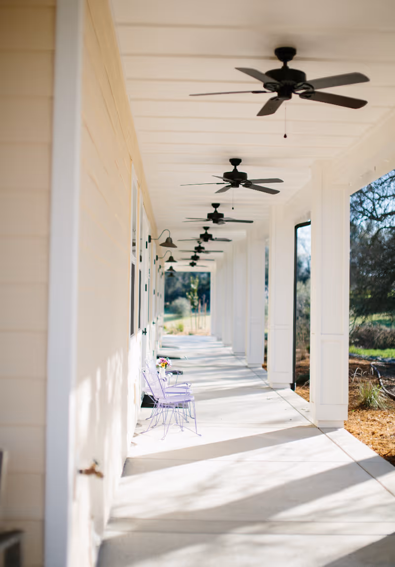 A long covered outdoor walkway with white columns, ceiling fans, and two purple chairs beside a small table with flowers.