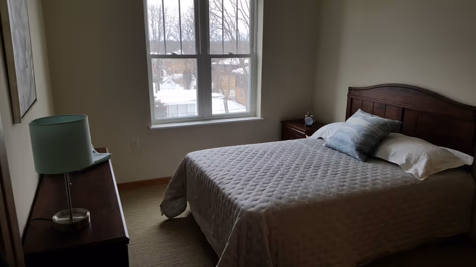 A bedroom with a wooden bed frame, a neatly made bed with a quilted gray bedspread, two pillows including one decorative pillow, a wooden nightstand with an alarm clock, a wooden dresser with a green lamp, and a window showing a snowy outdoor scene.