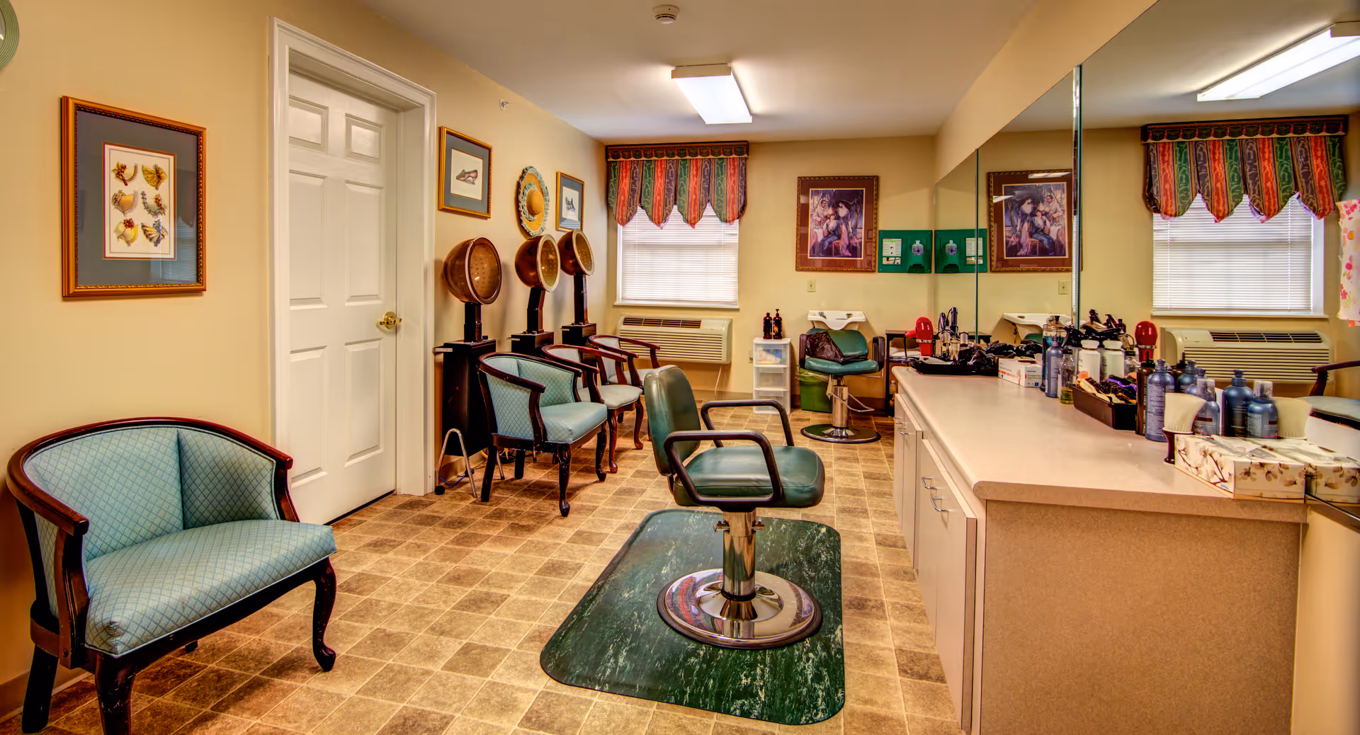 Interior of a salon room with a green salon chair in the center on a protective mat, a counter with various hair care products and tools, three vintage hair dryers with chairs underneath, framed artwork on the walls, two windows with colorful valances, and additional seating chairs along the walls.