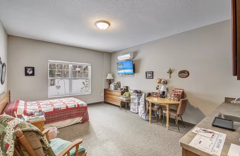 Bright senior studio room with a single bed, dresser, wall-mounted TV, small dining table, and a kitchenette counter.