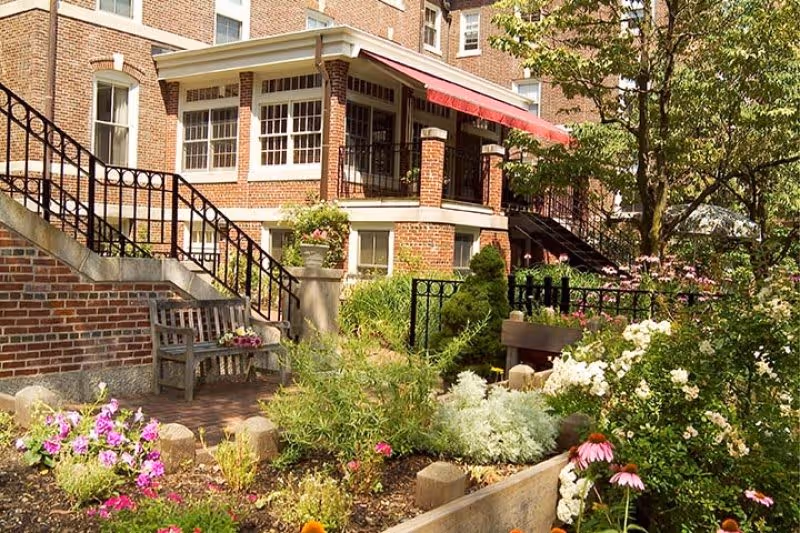 A garden area with colorful flowers and greenery in front of a brick building with large windows and a red awning. There are benches and black metal railings along the stairs leading up to the building entrance.