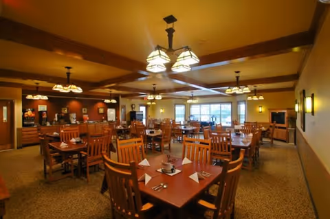 Well-lit dining room with multiple wooden tables and chairs set for meals in an assisted living community.