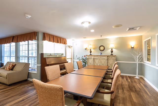 A well-lit interior room with a long rectangular table surrounded by eight cushioned chairs. To the left, there is a beige sofa near large windows with brown valances. Against the far wall, there is a wooden sideboard with two lamps, a clock, and decorative items. The floor is wooden, and the walls are painted light green with white trim.
