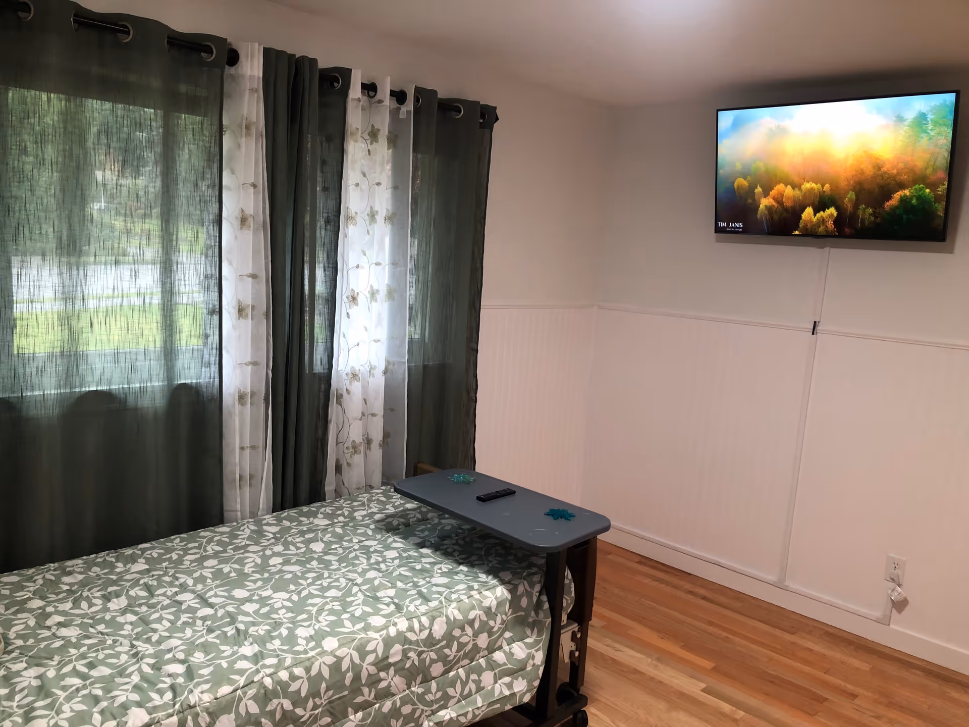 Small private bedroom with a single bed covered in green floral bedding, dark green curtains, a wall-mounted TV, and an overbed table.