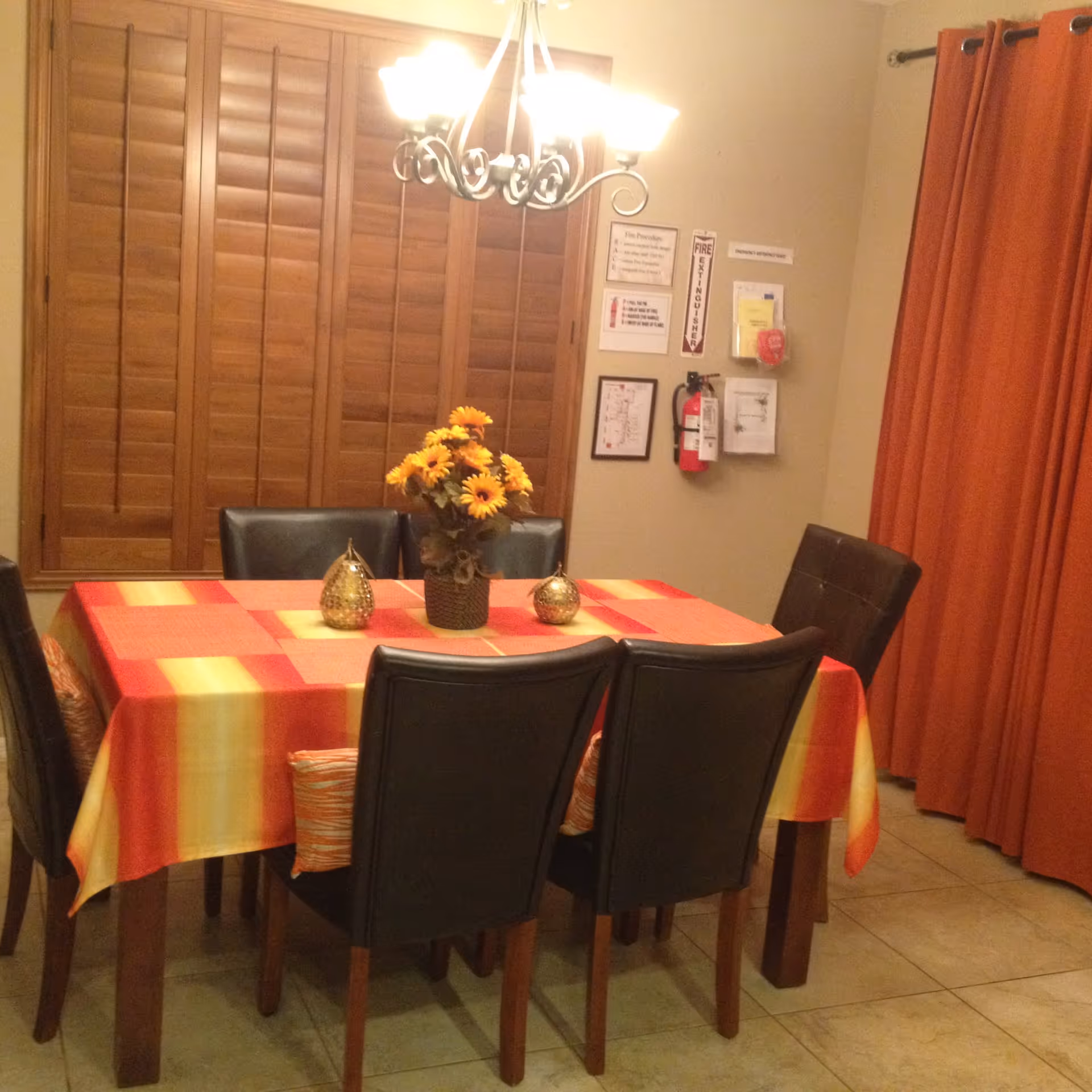 Dining area with a table covered by an orange and yellow tablecloth, six chairs, a sunflower centerpiece, chandelier, wooden shutters and orange curtains.