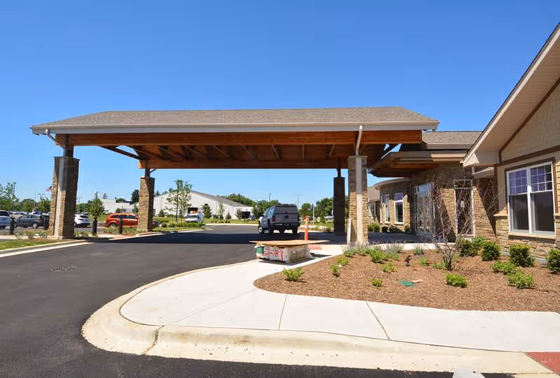 Entrance area of Silverado St. Charles Memory Care Community featuring a covered drop-off zone with wooden beams and stone pillars, surrounded by landscaped plants and a paved driveway under a clear blue sky.