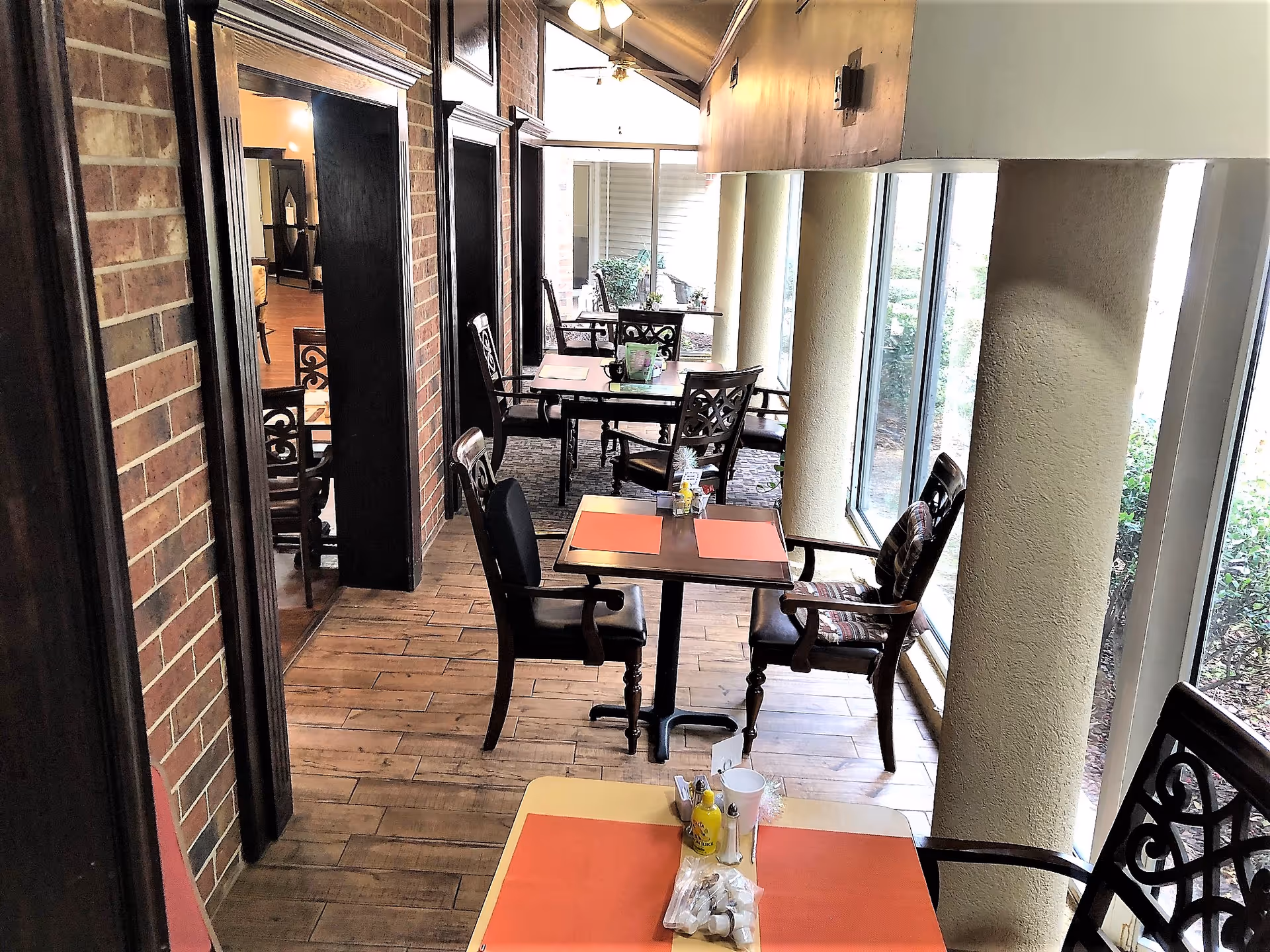 Indoor dining area with multiple tables and chairs arranged along large windows. Each table has orange placemats and condiments. The floor is wooden, and the walls have a brick and beige finish. The space is well-lit with natural light coming through the windows.
