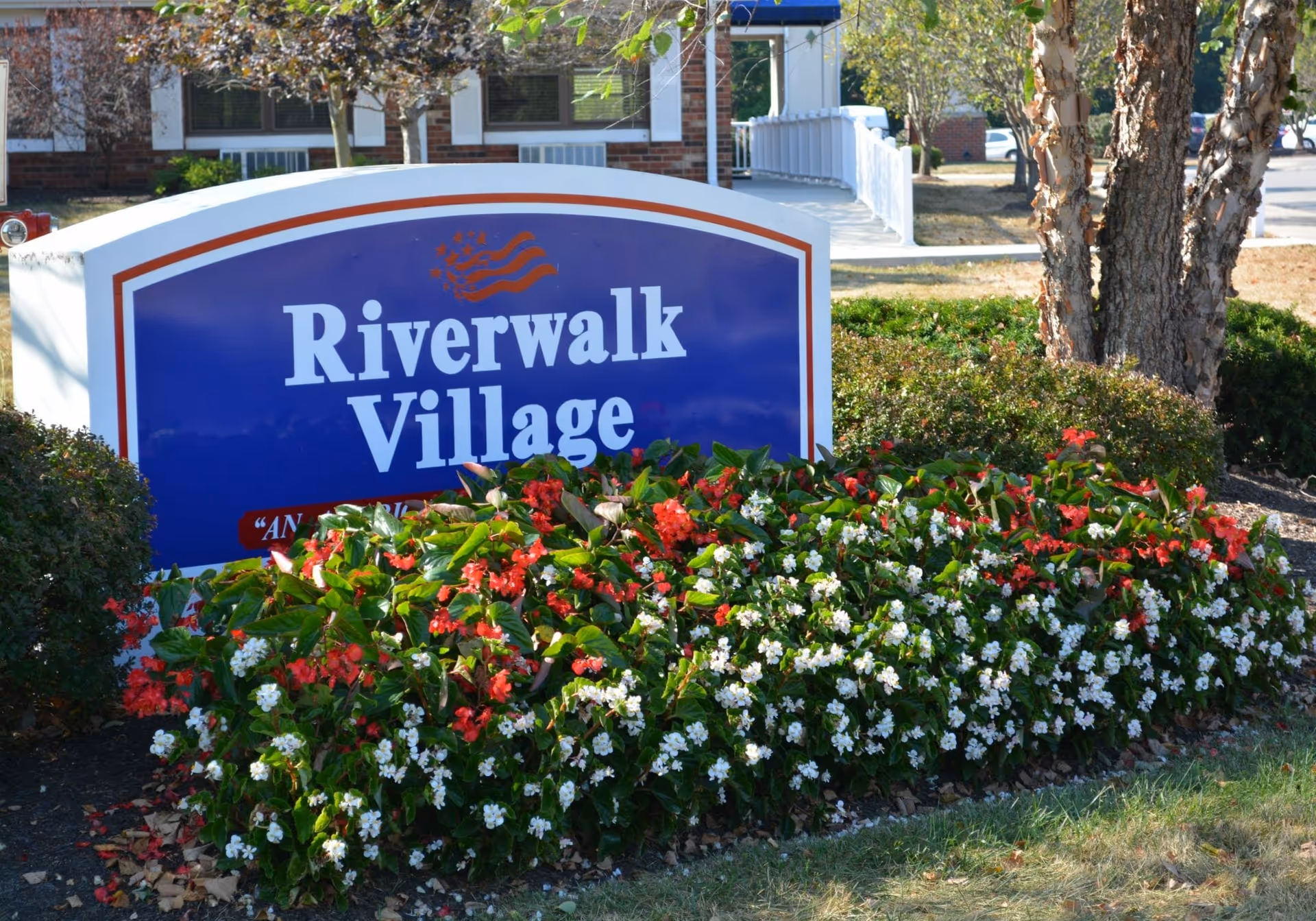 A blue and white sign for Riverwalk Village surrounded by red and white flowers and greenery, with a building and trees in the background.