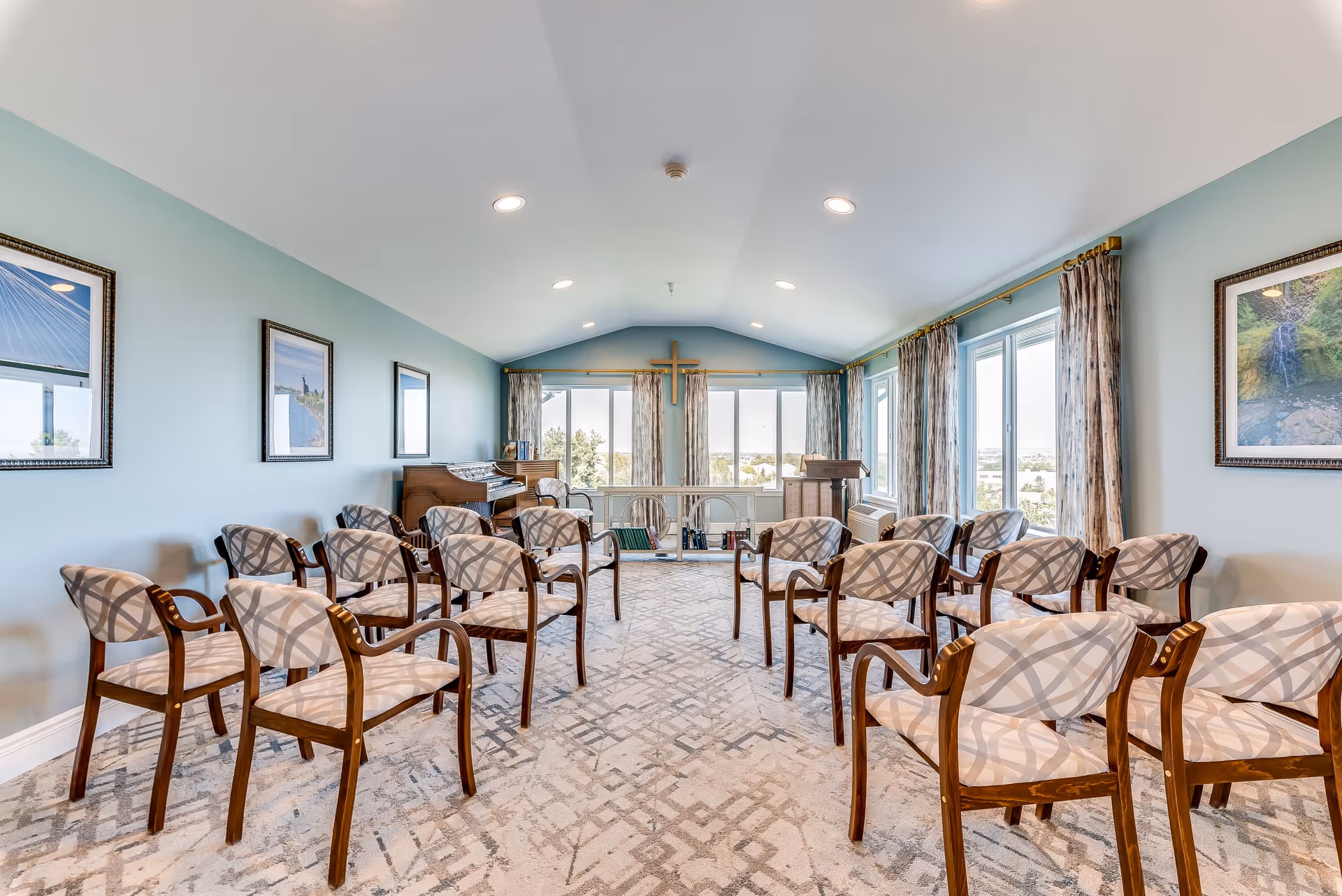 A bright room with multiple wooden chairs arranged in rows facing a small stage area with a wooden podium and a piano. The room has large windows with curtains, light blue walls, framed pictures, and a cross mounted on the wall above the stage area.