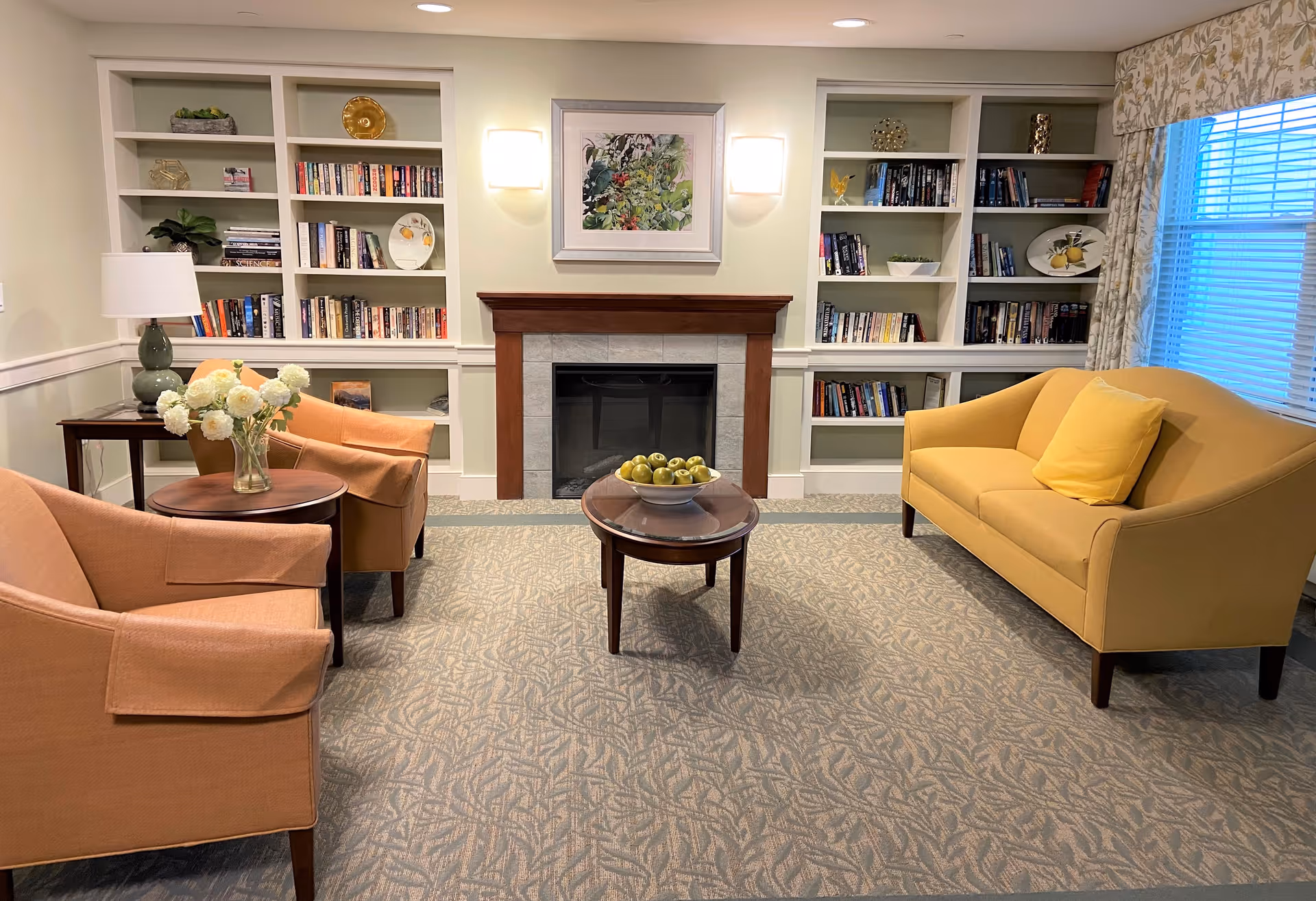 A cozy living room with a yellow couch and two peach armchairs arranged around a small wooden coffee table with a bowl of green apples. Behind the seating area is a fireplace with a wooden mantle, flanked by built-in bookshelves filled with books and decorative items. A framed floral painting hangs above the fireplace, and a window with patterned curtains is on the right side of the room.