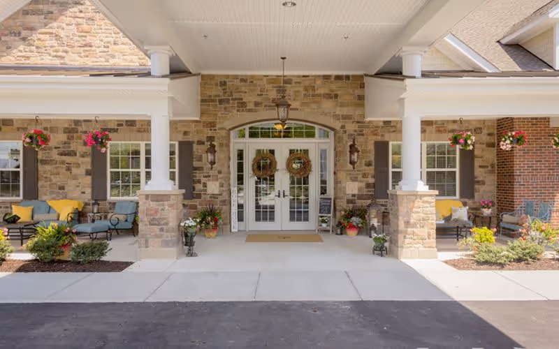 Front entrance of a senior living facility with stone and brick exterior walls, double glass doors decorated with wreaths, white columns supporting a covered porch, hanging flower baskets, and outdoor seating areas with chairs and cushions on either side.
