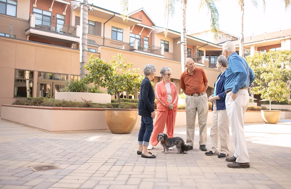 Five elderly people standing and chatting outdoors in a courtyard area of a retirement community with a small dog on a leash. The background shows a multi-story building with balconies, palm trees, and large potted plants.