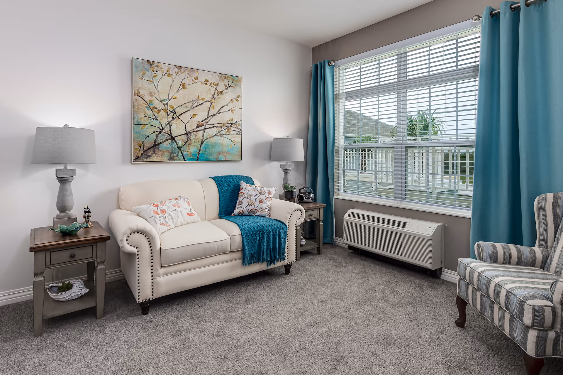 A cozy living room with a cream-colored loveseat adorned with floral pillows and a teal throw blanket. There are two matching side tables with gray lamps on either side of the loveseat. A large window with teal curtains lets in natural light, and a striped armchair is positioned to the right. The walls are light-colored, and a painting of tree branches with birds hangs above the loveseat. The floor is carpeted in gray.