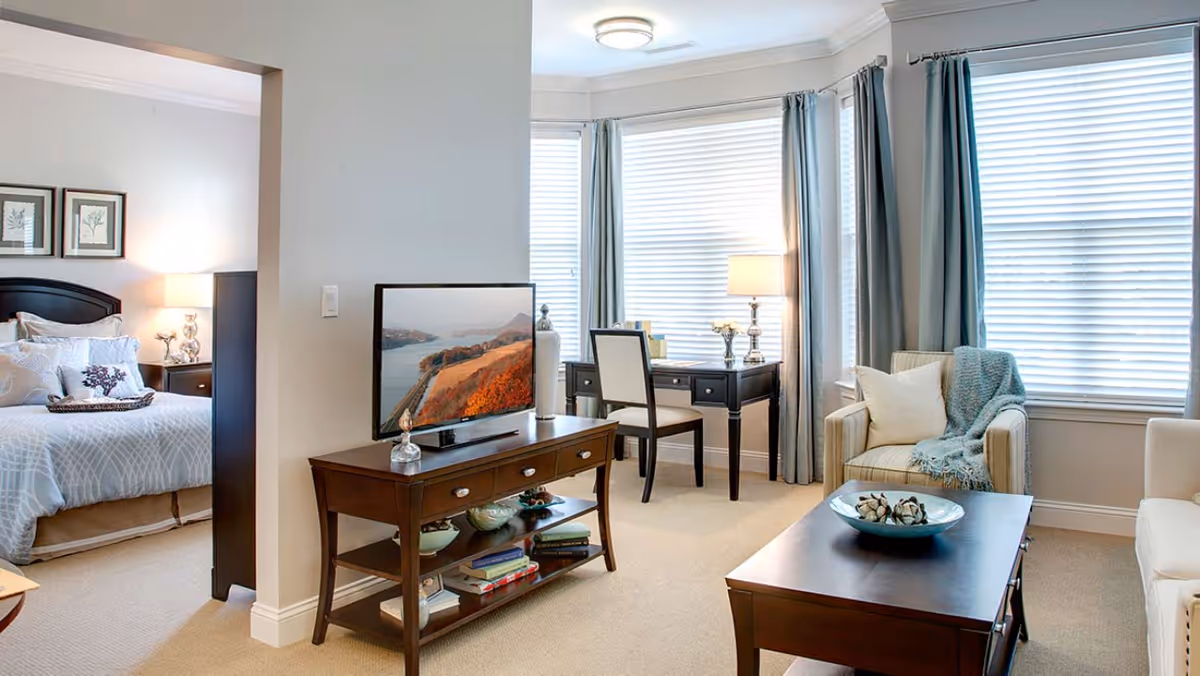 Bright open living room with a TV console, coffee table, armchair and desk, with a view into a bedroom.