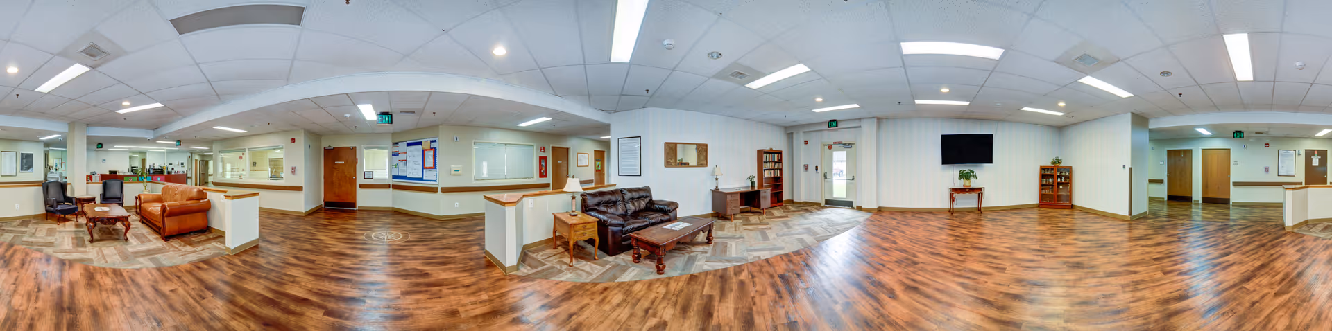 Panoramic view of a senior living facility common area with wood flooring, white walls, and recessed lighting. The space includes seating areas with brown and black leather sofas, wooden coffee tables, a TV mounted on the wall, and a few wooden cabinets with plants and decorations. There are multiple doors and windows along the walls, and a reception or service window area is visible.