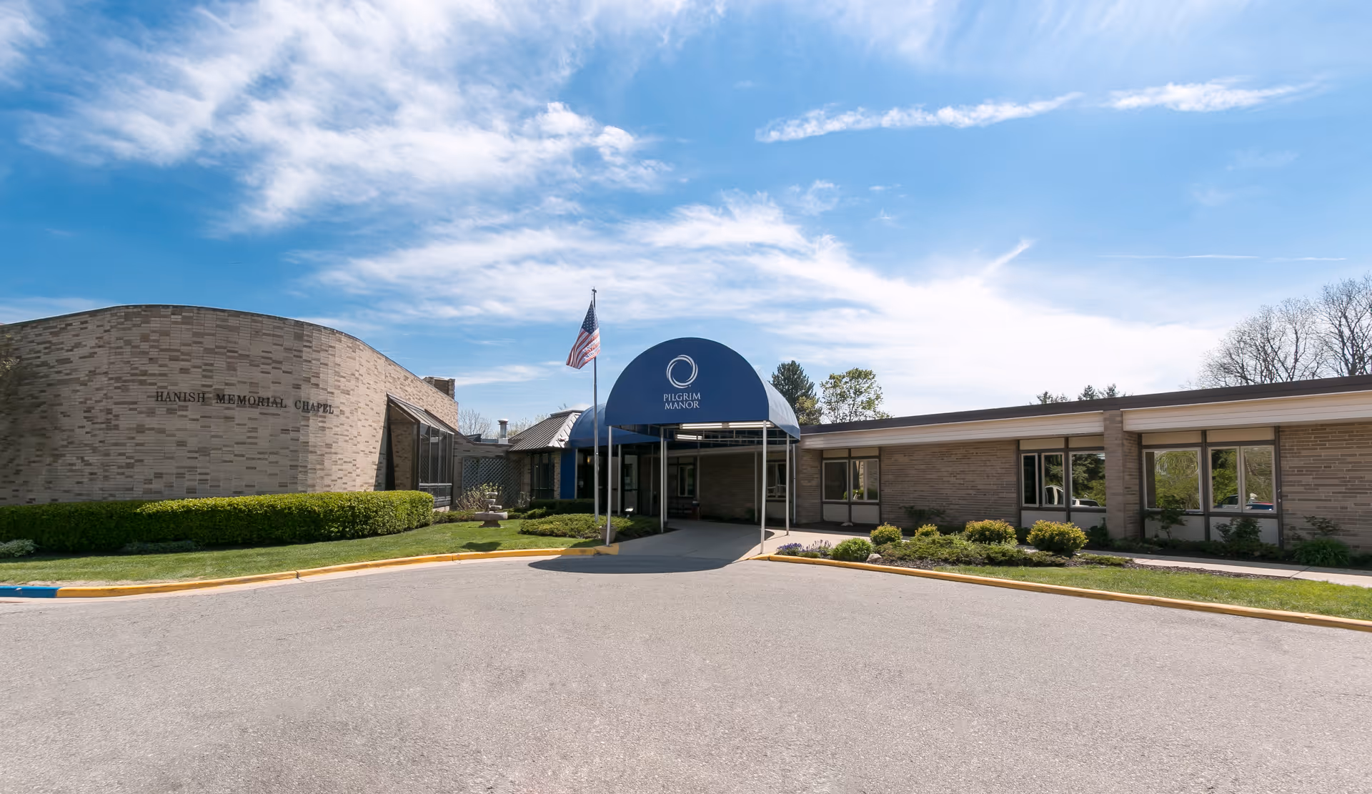 Exterior front view of a single-story brick building with a blue awning entrance labeled Pilgrim Manor, an American flag on a flagpole, and a curved section labeled Hanish Memorial Chapel, under a partly cloudy blue sky.