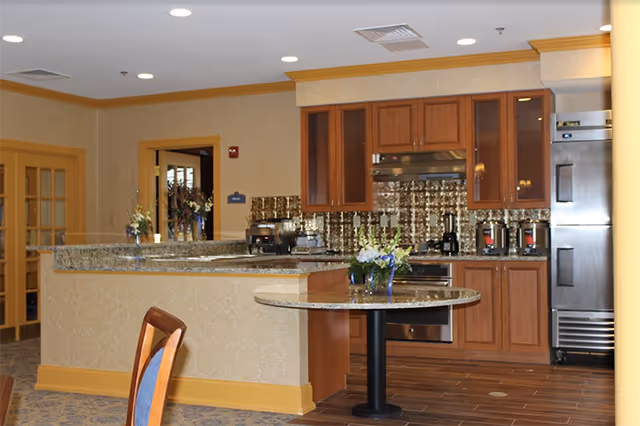 Interior view of a kitchen area in an assisted living facility featuring wooden cabinets, a stainless steel refrigerator, oven, and various kitchen appliances on the countertop. There is a round table with a flower arrangement in the foreground and a partial view of a chair. The walls have a light yellow patterned wallpaper and the ceiling has recessed lighting.