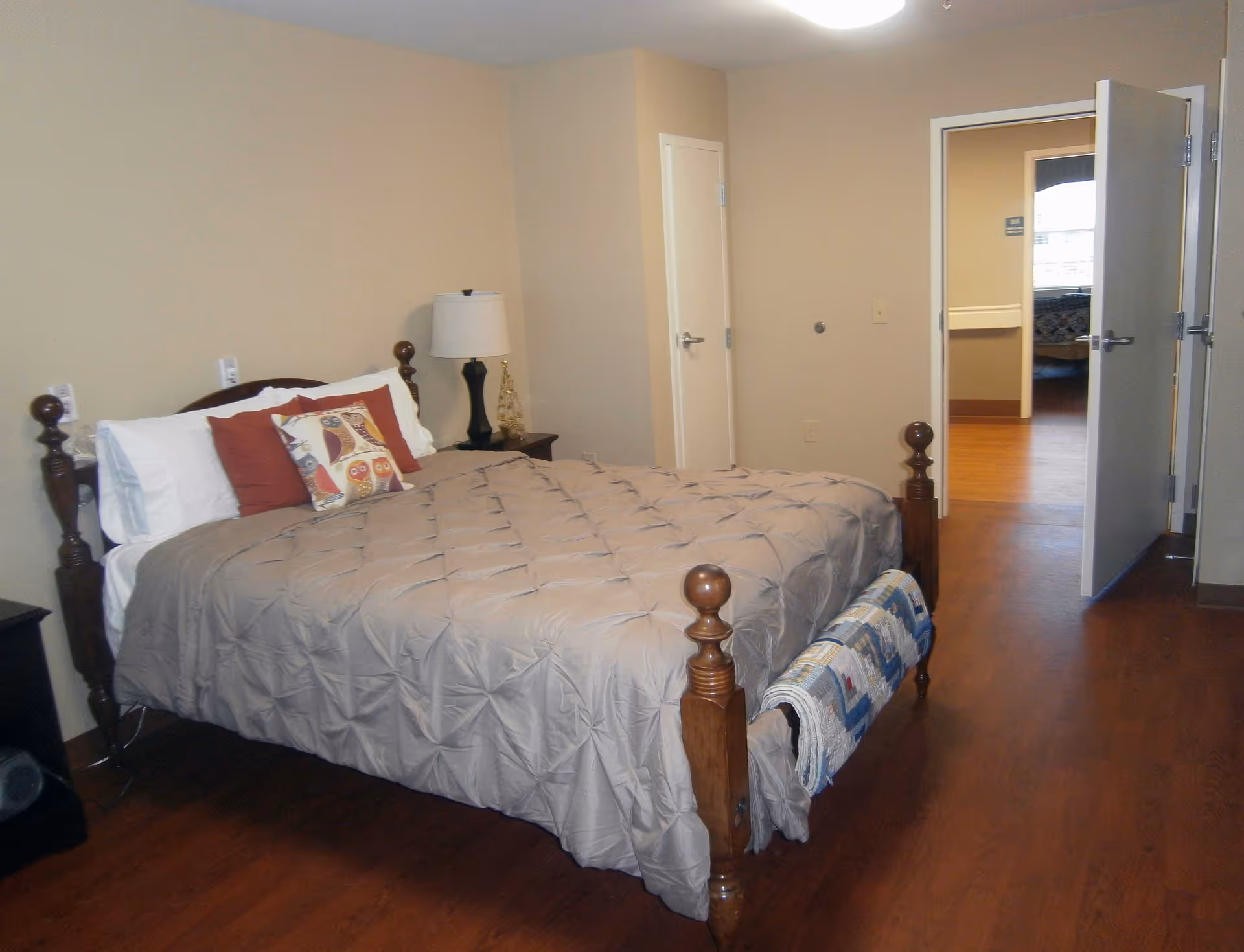 A bedroom with a wooden bed frame, beige comforter, and decorative pillows including one with an owl design. There is a nightstand with a lamp and decorative item next to the bed. The room has wooden flooring and beige walls. An open door reveals another room with similar flooring and a window.