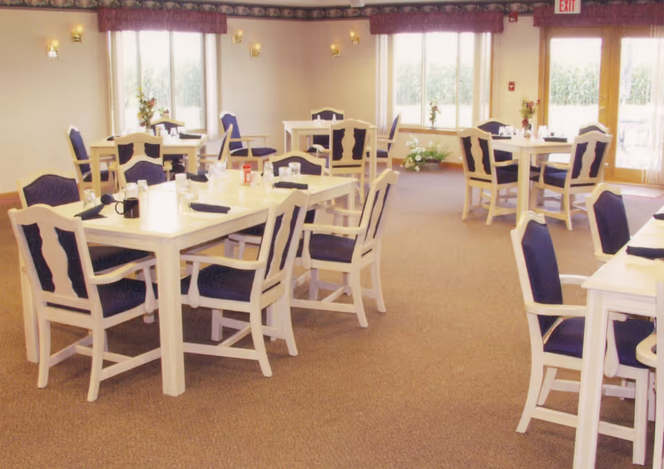 Bright senior dining room with multiple white wooden tables and blue-upholstered chairs set for meals.