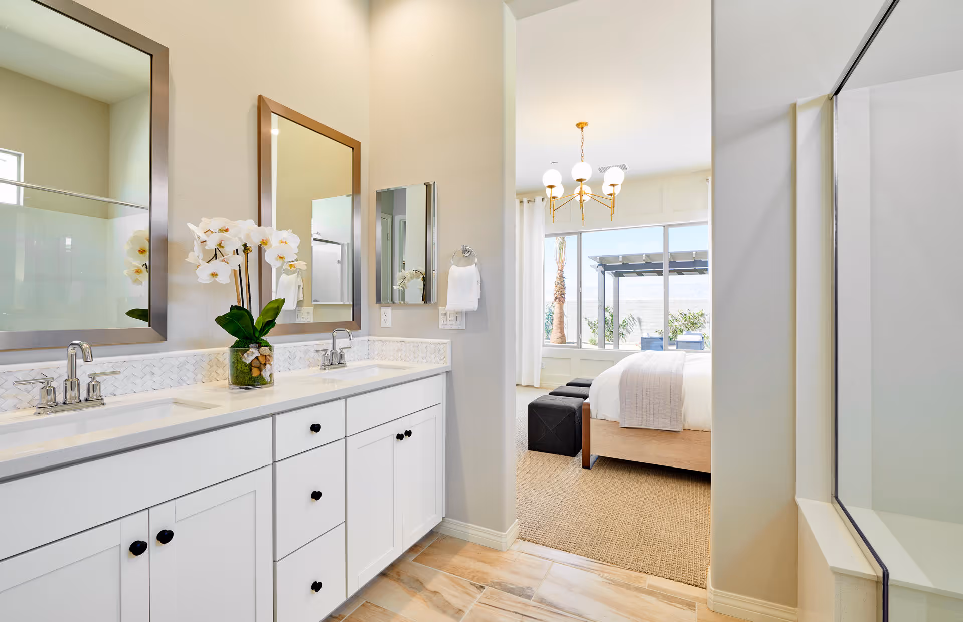 Bright bathroom with double sinks, white cabinetry, and two large mirrors above the sinks. A vase with white orchids is placed on the countertop. The bathroom opens into a bedroom with a bed, two black ottomans, a large window, and a chandelier. The bedroom has a view of an outdoor patio area with palm trees.