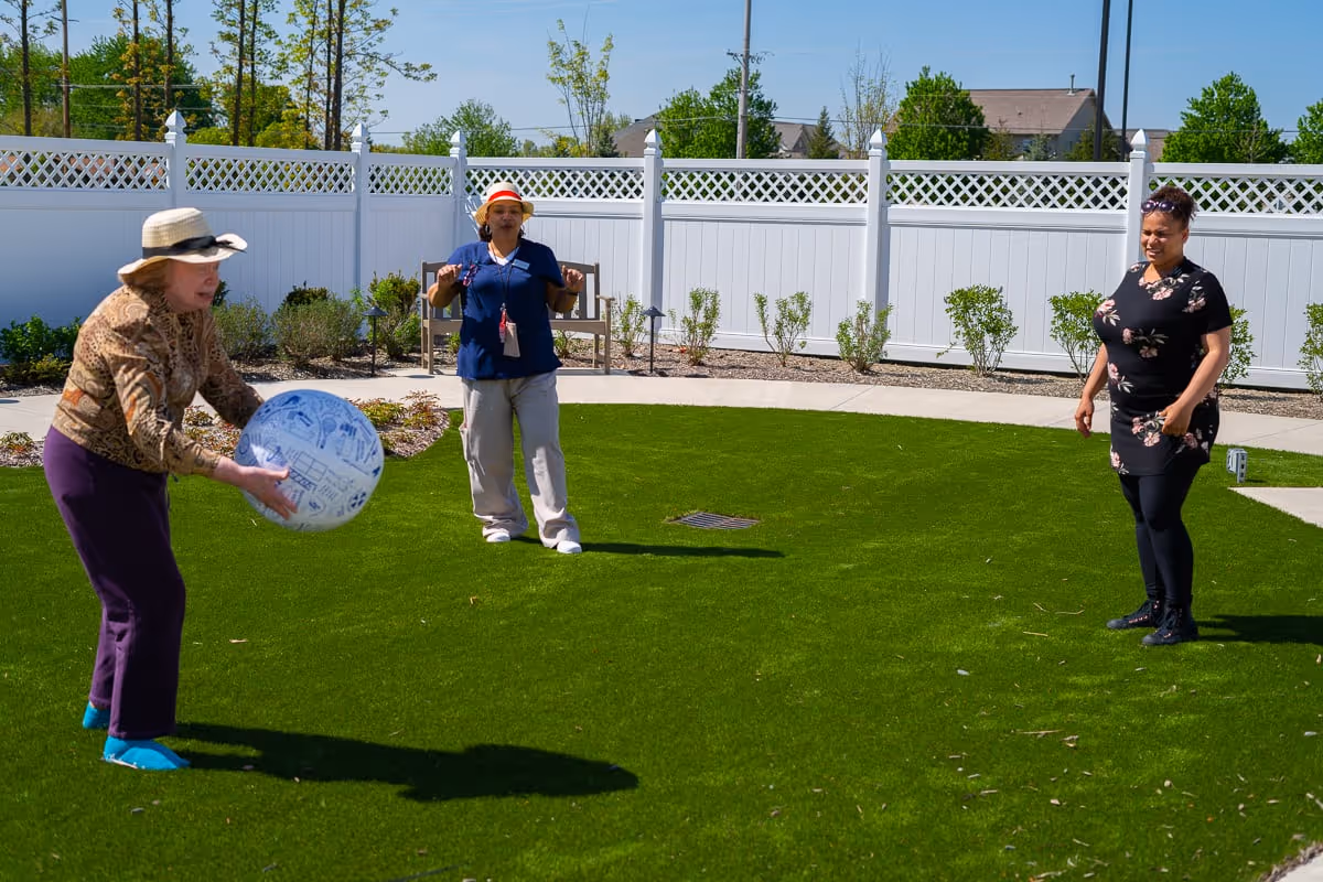 Three women standing on a green lawn enclosed by a white fence. One woman is holding a large blue and white ball, while the other two women watch and smile. The setting appears to be an outdoor recreational area with some plants and a clear blue sky.