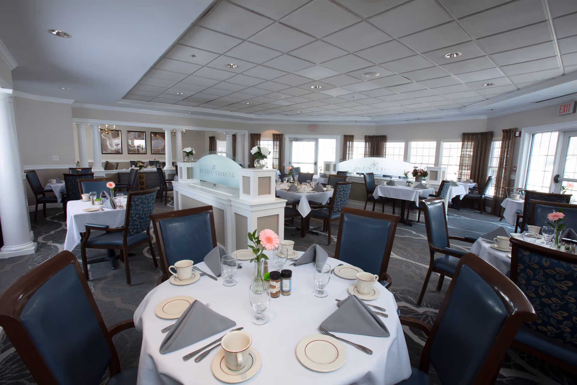 A bright and spacious dining room at Heights Crossing Assisted Living, featuring round and rectangular tables set with white tablecloths, gray napkins, cups, plates, and glassware. Each table has a small vase with a pink flower. The room has large windows with brown curtains, white columns, and a carpeted floor with a floral pattern.